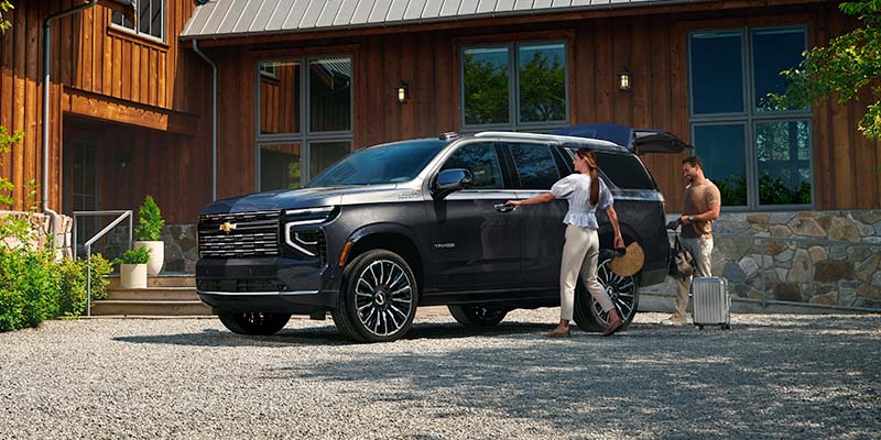 A couple standing beside a Chevrolet Tahoe, packing luggage for a trip in front of a modern home.
