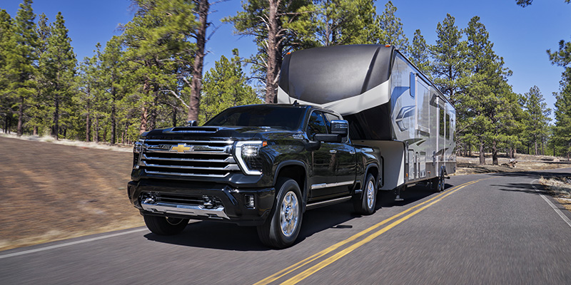 Black Chevrolet Silverado 1500 towing a large fifth-wheel trailer on a paved road surrounded by tall pine trees