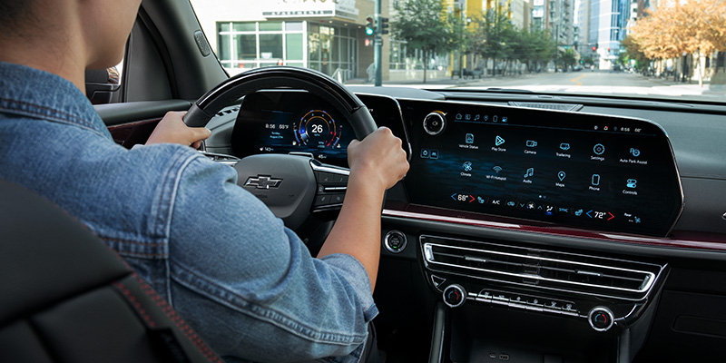 Person driving a Chevrolet vehicle, showing the steering wheel, digital instrument cluster, and large central touchscreen display with navigation and infotainment features