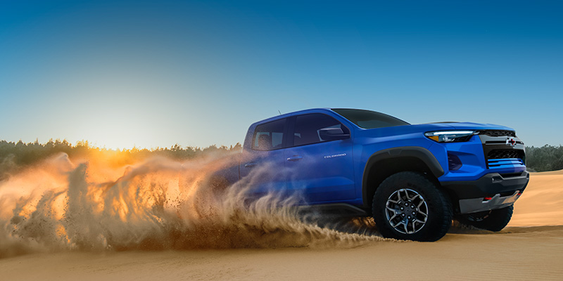 Blue pickup truck drifting across a sandy desert landscape, kicking up a large cloud of dust under a clear sky at sunset.