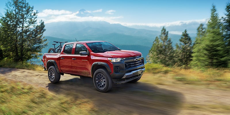 Red Chevrolet pickup truck driving on a forest trail with mountains in the background.
